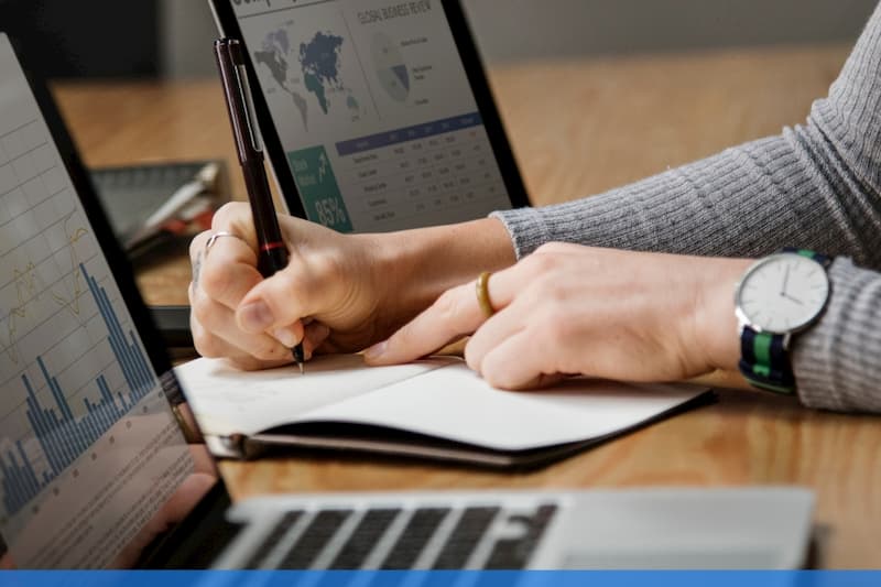 Woman writing on paper between two laptops showing data